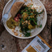 Plate of food with a fork, rice, greens, and a packet of GH Produce curry mix on a stone surface.